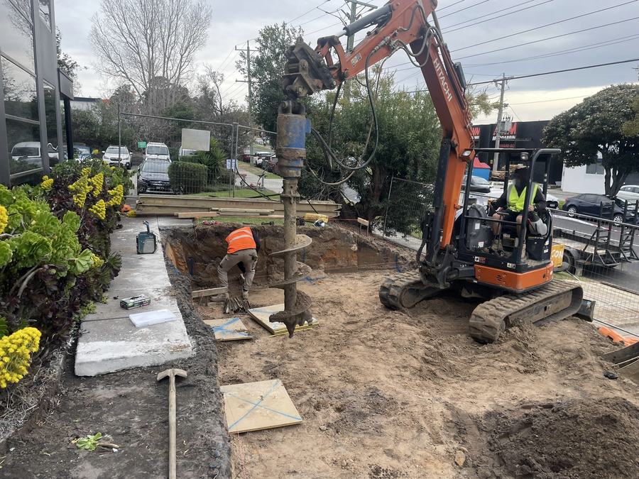 Excavator with auger attachment drilling footings for a failed retaining wall replacement